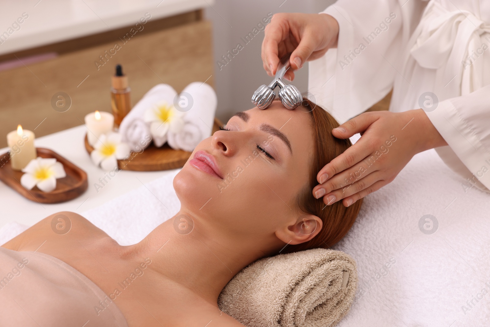 Photo of Young woman receiving facial massage with metal roller in beauty salon, closeup