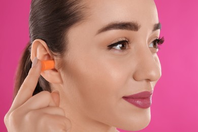 Young woman inserting foam ear plug on pink background, closeup