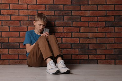 Photo of Boy with smartphone sitting on floor near brick wall, space for text. Children's bullying
