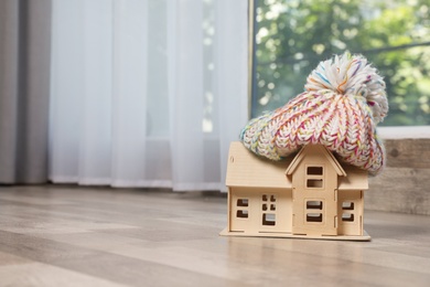 Photo of Wooden house model in hat on floor indoors, space for text. Heating efficiency