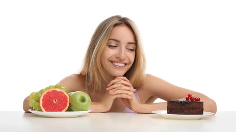 Woman choosing between cake and healthy fruits at table on white background