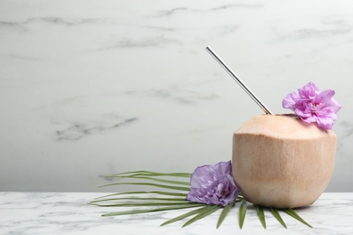 Photo of Young peeled coconut with straw, palm leaf and flowers on white marble table. Space for text