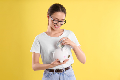 Young woman putting money into piggy bank on yellow background