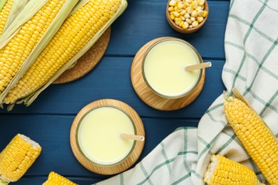 Photo of Tasty fresh corn milk in glasses and cobs on blue wooden table, flat lay