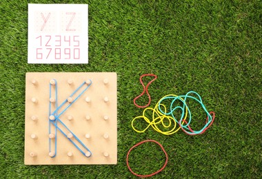 Wooden geoboard with letter K made of rubber bands and activity book on artificial grass, flat lay. Educational toy for motor skills development