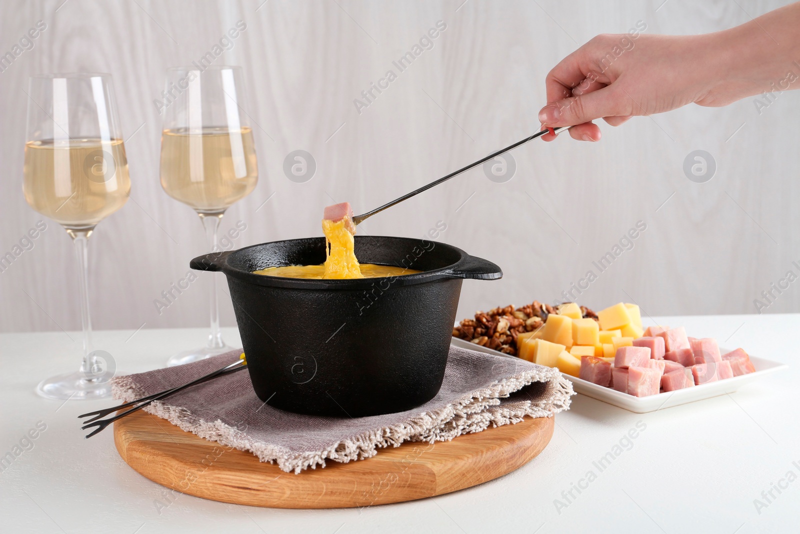 Photo of Dipping piece of ham into fondue pot with tasty melted cheese at white table, closeup