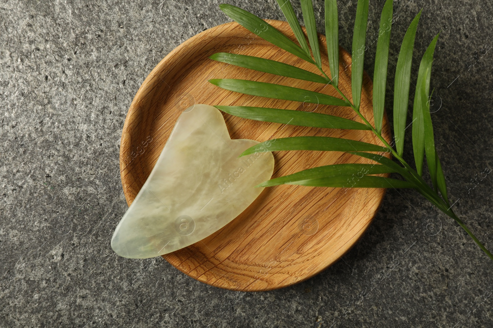 Photo of Jade gua sha tool and green leaf on grey table, flat lay