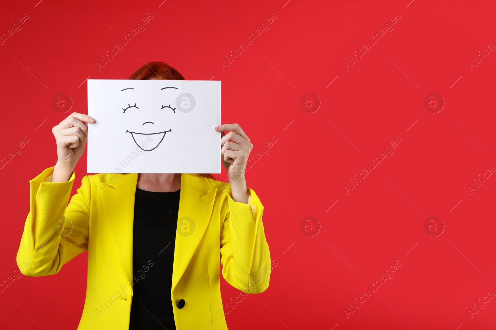 Photo of Woman hiding behind sheet of paper with happy face on red background, space for text