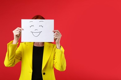 Woman hiding behind sheet of paper with happy face on red background, space for text