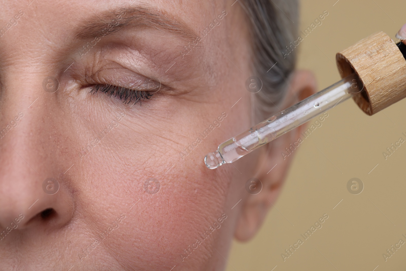 Photo of Senior woman applying cosmetic product on her aging skin against beige background, closeup. Rejuvenation treatment