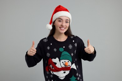 Photo of Happy young woman in Christmas sweater and Santa hat showing thumbs up on grey background