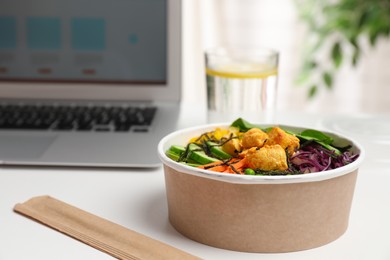 Photo of Delicious salad with chicken and vegetables in paper bowl near laptop on white table. Space for text