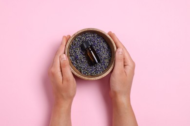 Woman holding bowl with lavender essential oil and flowers on pink background, top view