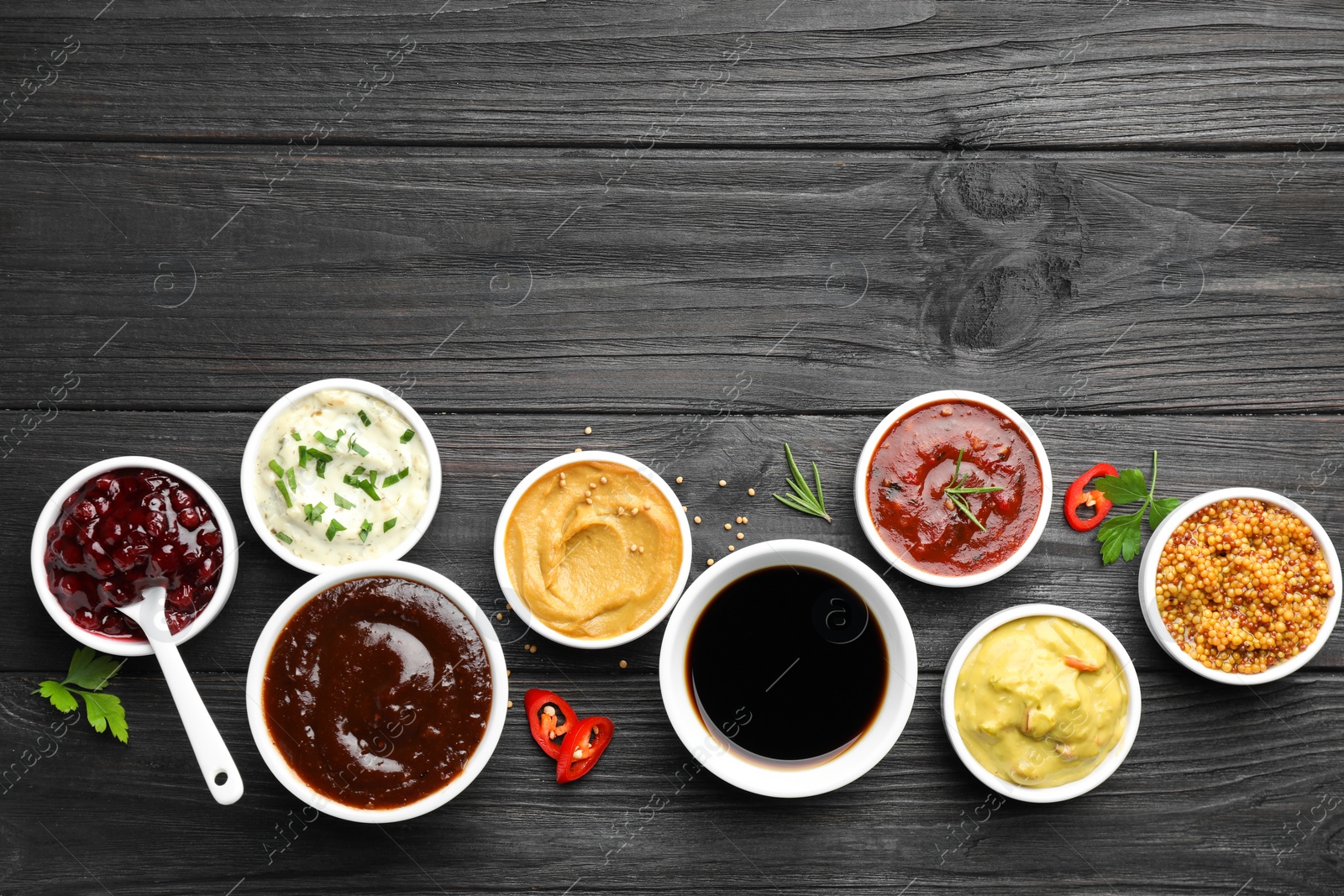 Photo of Different tasty sauces in bowls, parsley, chili pepper and rosemary on black wooden table, flat lay. Space for text