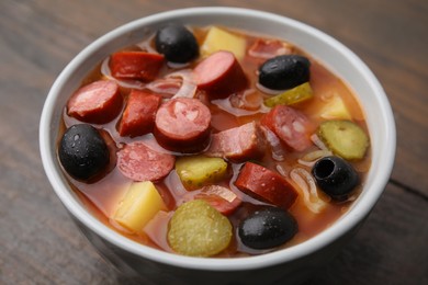 Photo of Meat solyanka soup with thin dry smoked sausages in bowl on wooden table, closeup