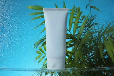 Photo of Tube with moisturizing cream and palm leaves on light blue background, view through wet glass