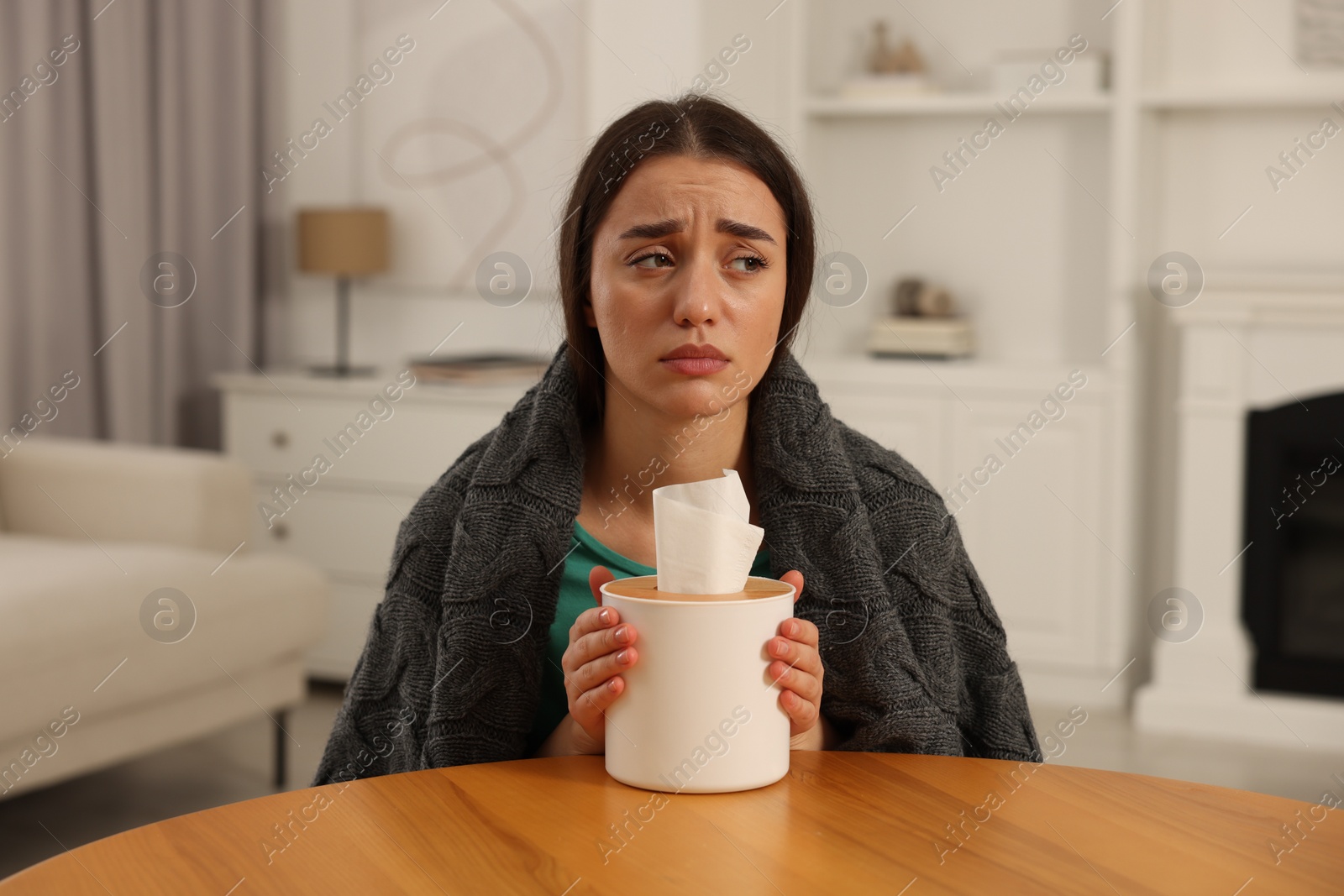 Photo of Sick woman wrapped in blanket with box of tissues at wooden table indoors. Cold symptoms