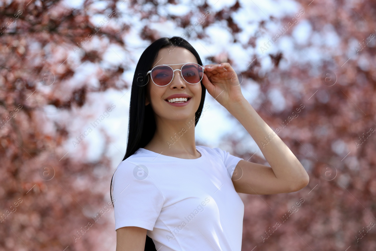 Photo of Pretty young woman with sunglasses near beautiful blossoming trees outdoors. Stylish spring look