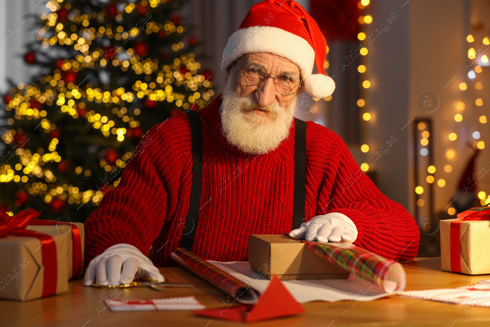 Photo of Santa Claus wrapping gift at his workplace in room decorated for Christmas