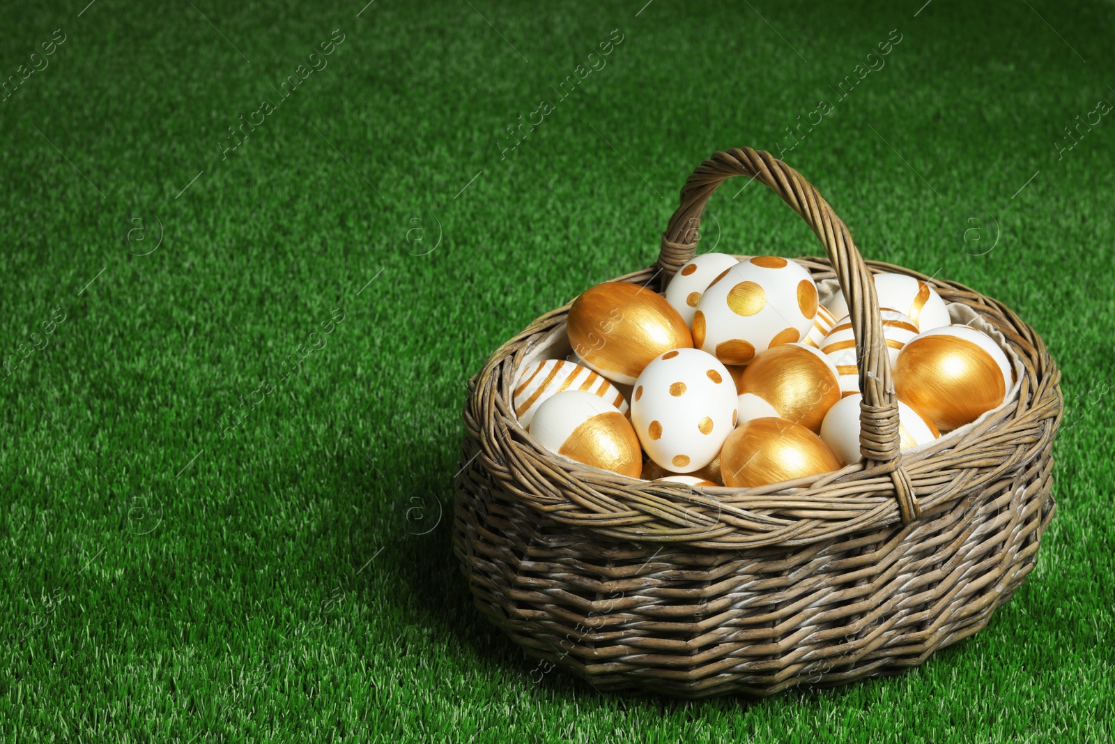 Photo of Wicker basket of traditional Easter eggs decorated with golden paint on green lawn, space for text