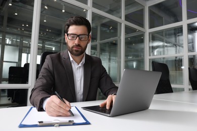 Photo of Man writing notes while working on laptop at white desk in office