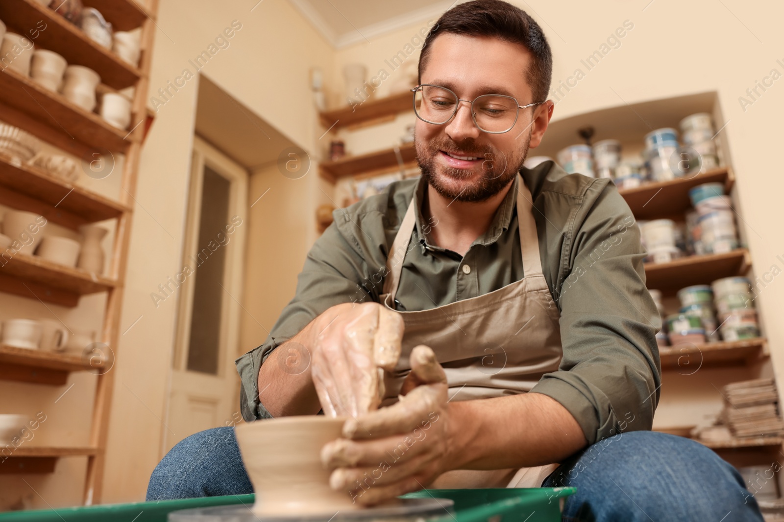 Photo of Clay crafting. Happy man making bowl on potter's wheel in workshop, low angle view