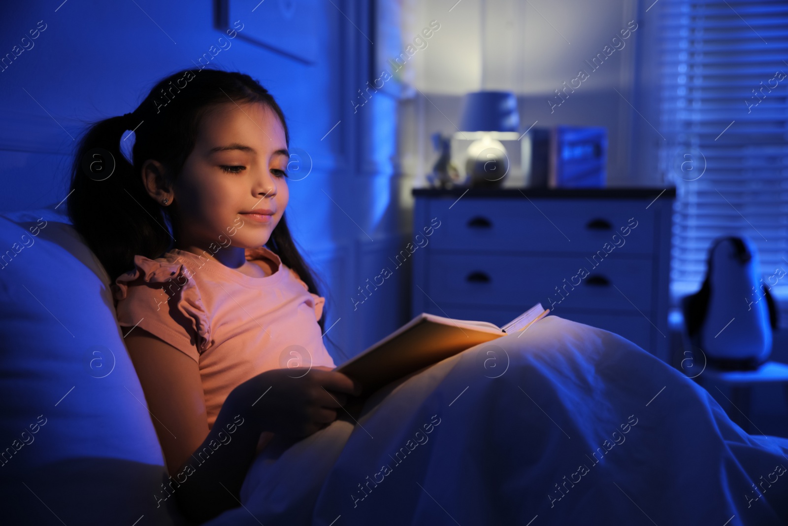 Photo of Little girl reading fairy tale in dark bedroom