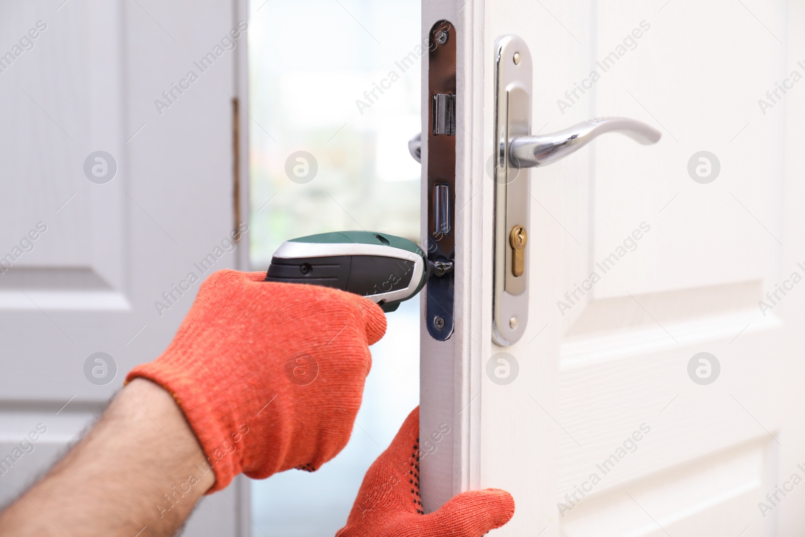 Photo of Handyman with screw gun repairing door lock indoors, closeup