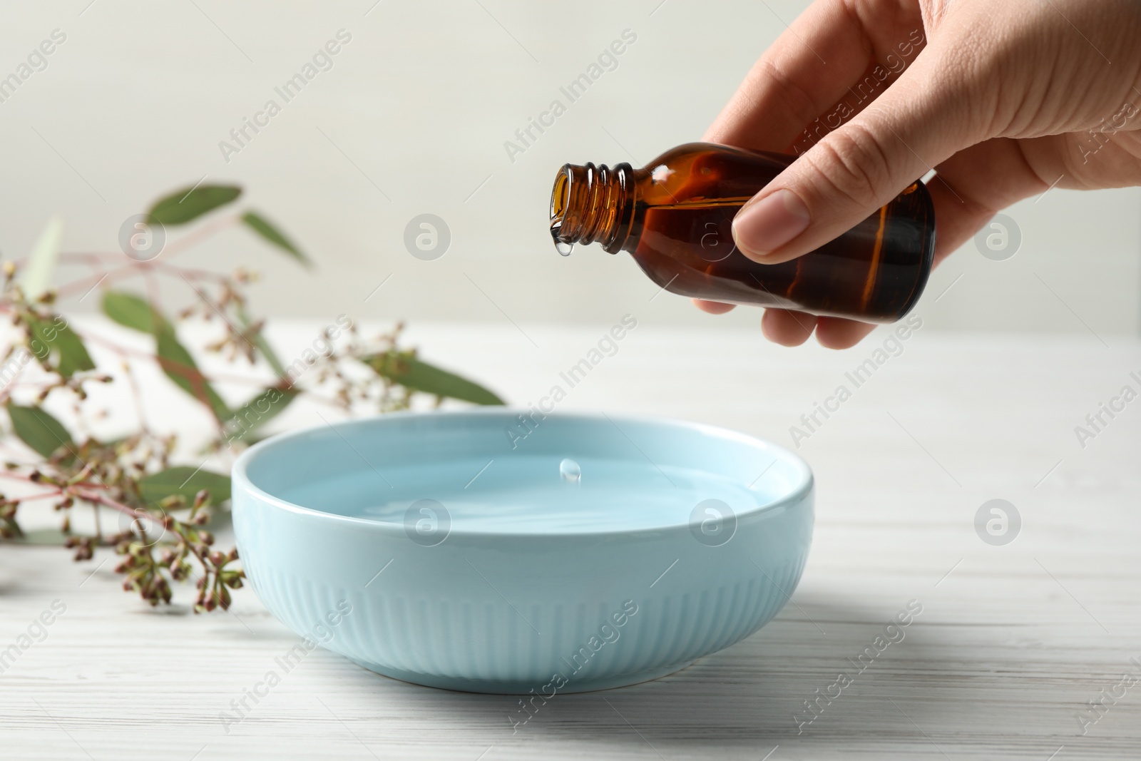 Photo of Woman dripping eucalyptus essential oil from bottle into bowl at white wooden table, closeup