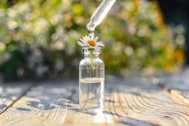 Dripping essential oil from pipette onto chamomile in bottle on white wooden table, closeup
