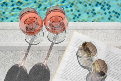 Glasses of tasty rose wine, open book and sunglasses on swimming pool edge