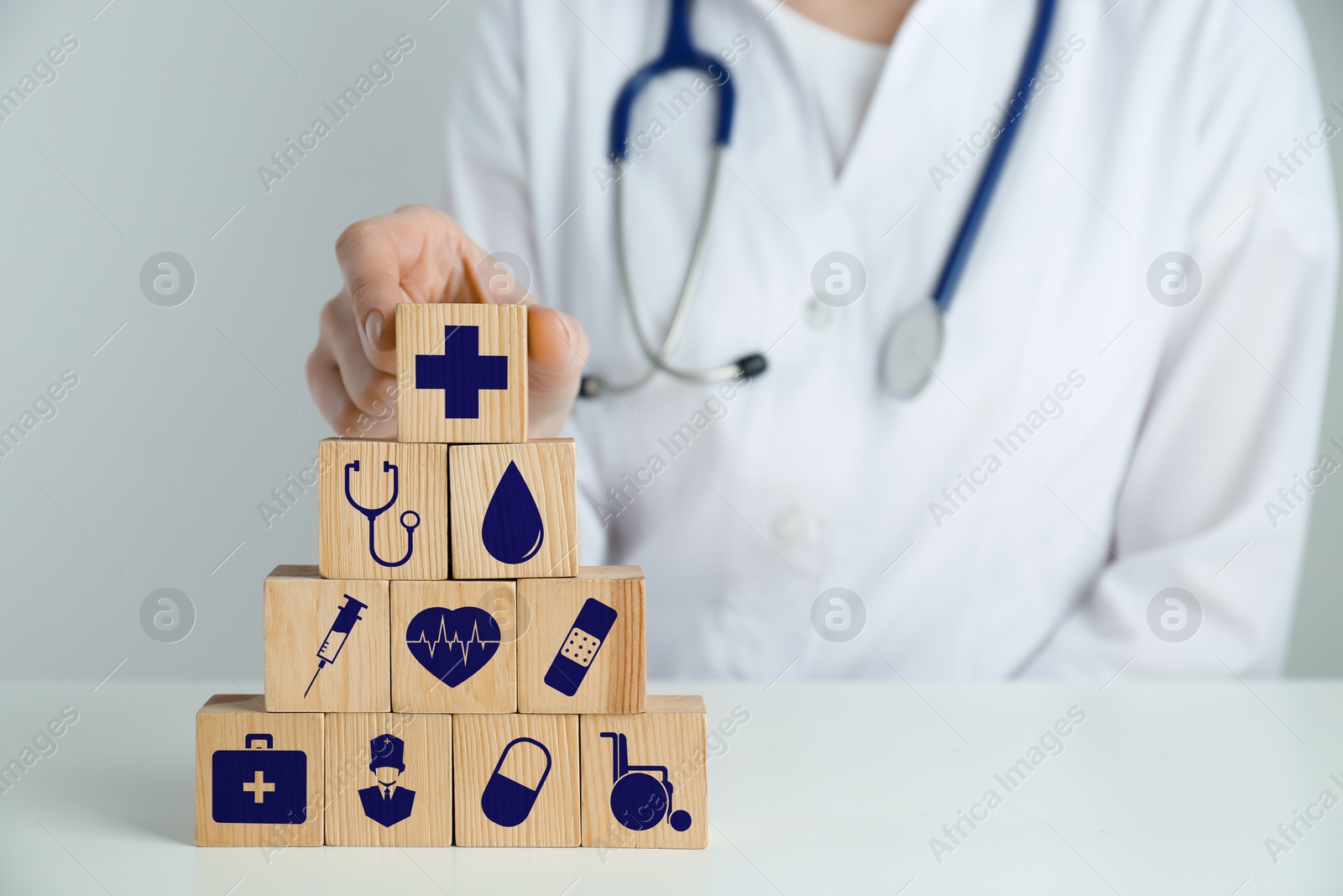 Image of Doctor building pyramid of wooden cubes with different icons at white table, closeup. Insurance concept