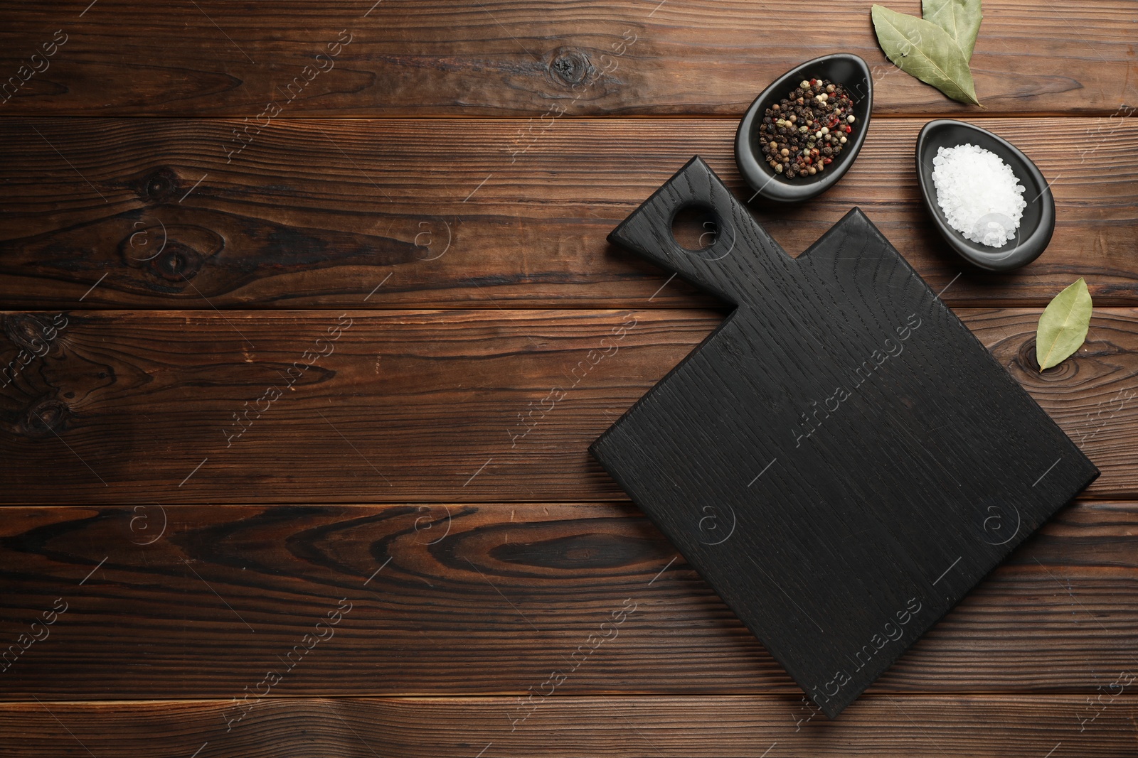 Photo of Black cutting board, salt, pepper and bay leaves on wooden table, flat lay. Space for text