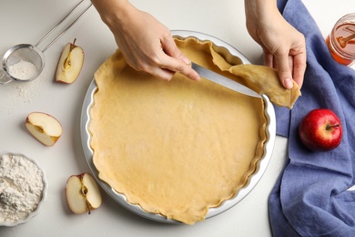 Photo of Woman cutting dough leftovers for traditional English apple pie in baking dish at white table, top view