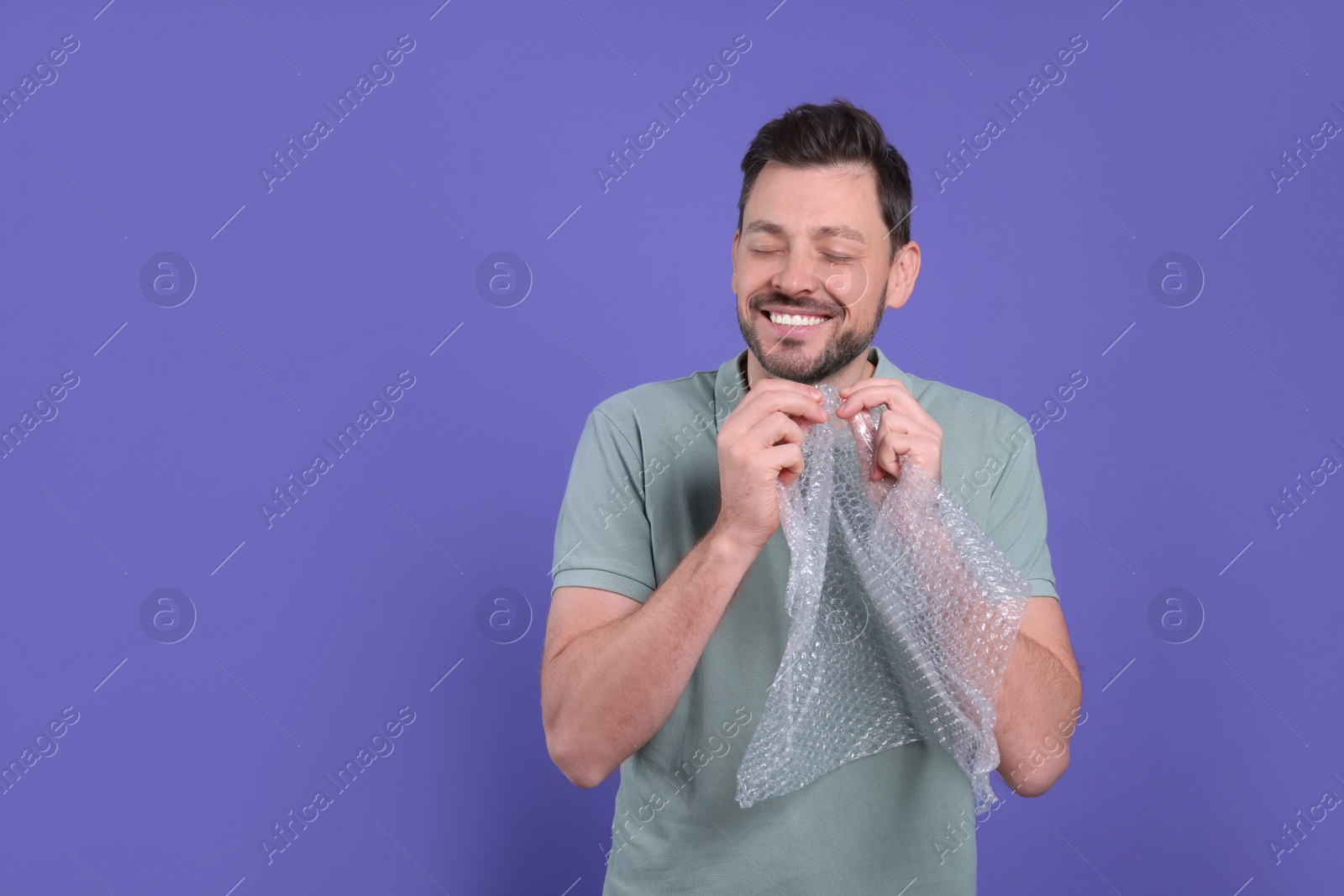 Photo of Happy man popping bubble wrap on purple background, space for text. Stress relief