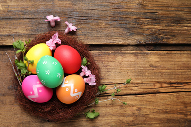 Photo of Colorful Easter eggs in decorative nest with flowers on wooden background, flat lay. Space for text