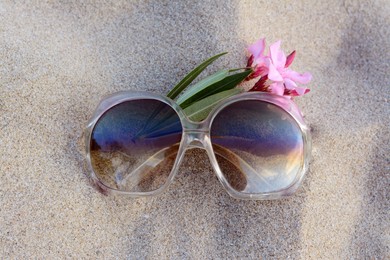 Beautiful sunglasses with tropical flower on sandy beach, top view