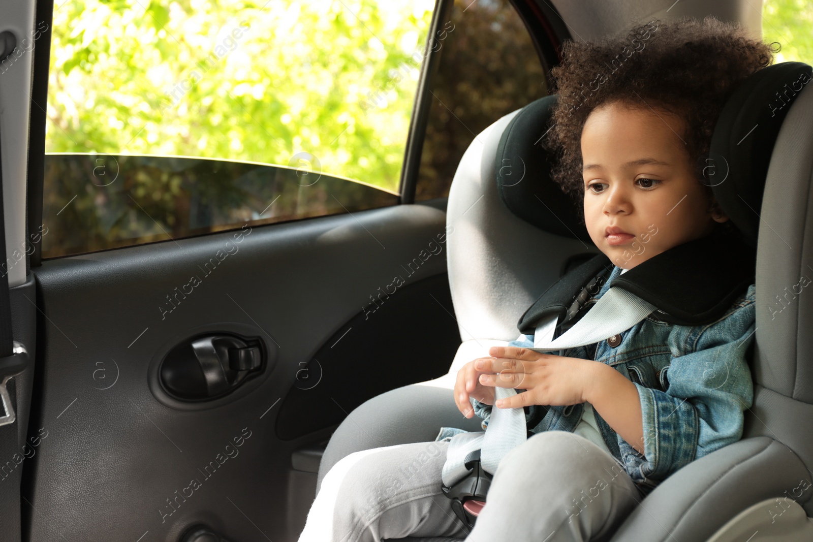 Photo of Cute African-American girl sitting in safety seat alone inside car. Child in danger