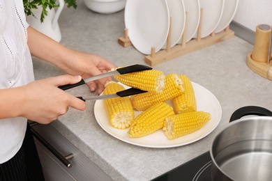 Photo of Woman taking corn cob from plate with tongs in kitchen, closeup