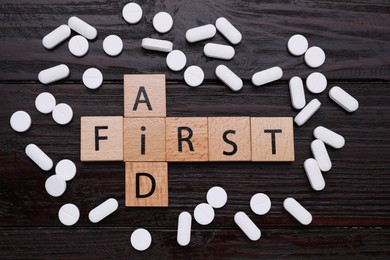 Photo of Words First Aid made of cubes and pills on black wooden table, flat lay