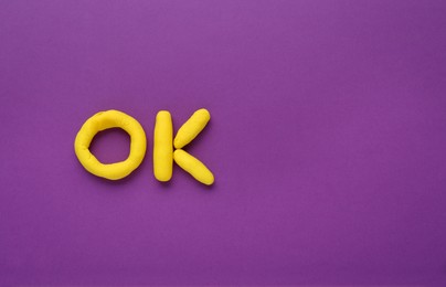 Photo of Word Ok made of yellow plasticine on purple background, top view. Space for text