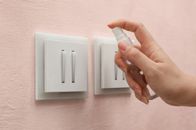 Photo of Woman spraying antiseptic onto light switch indoors, closeup