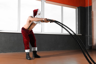 Photo of Young shirtless Santa Claus training with ropes in modern gym