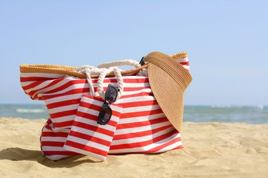 Stylish striped bag with visor cap and sunglasses on sandy beach near sea