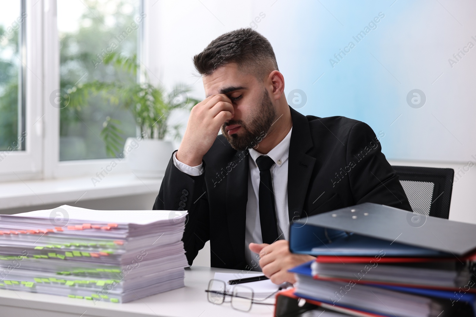 Photo of Overwhelmed man sitting at table with stacks of documents and folders in office