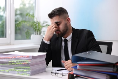 Overwhelmed man sitting at table with stacks of documents and folders in office