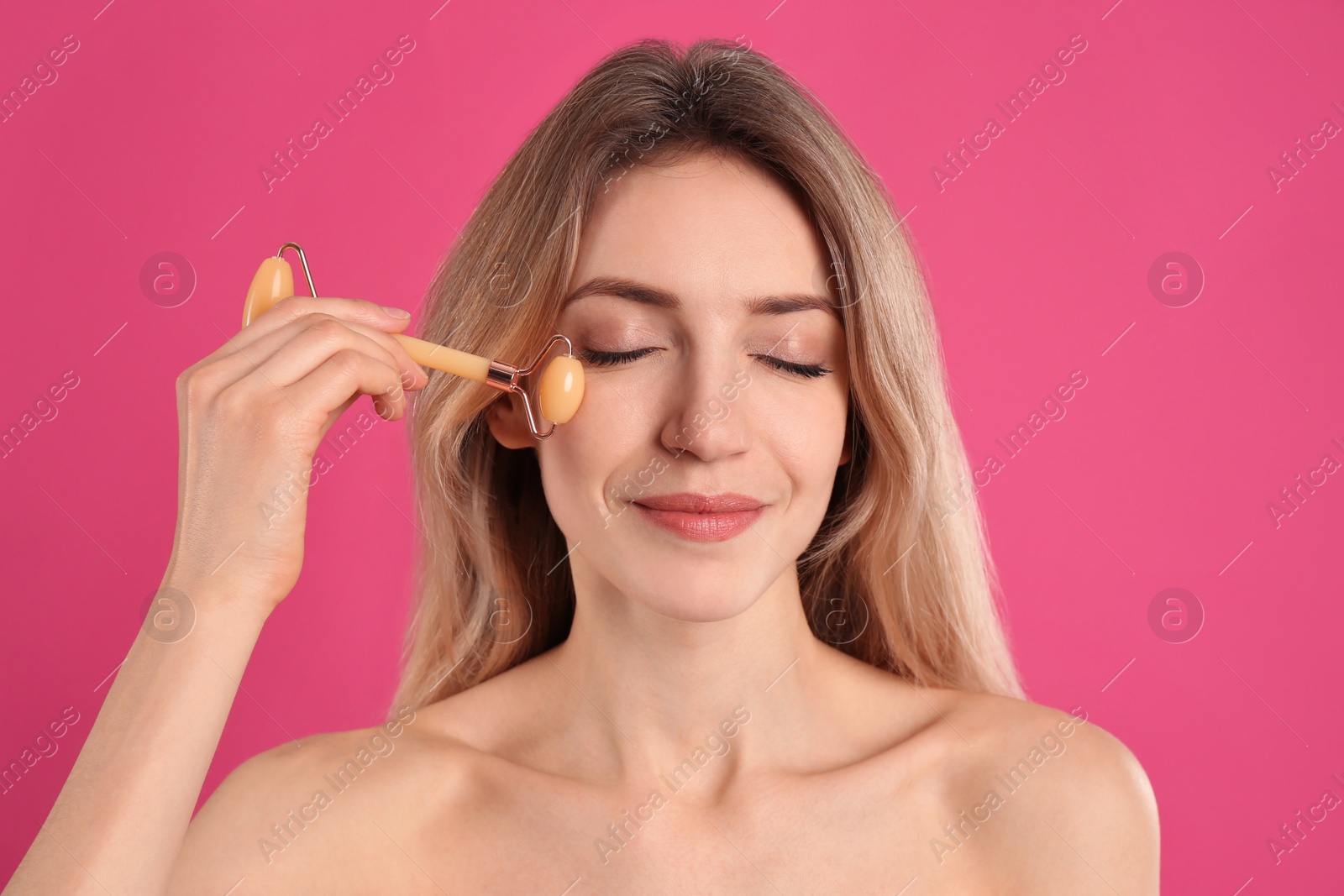 Photo of Young woman using natural jade face roller on pink background