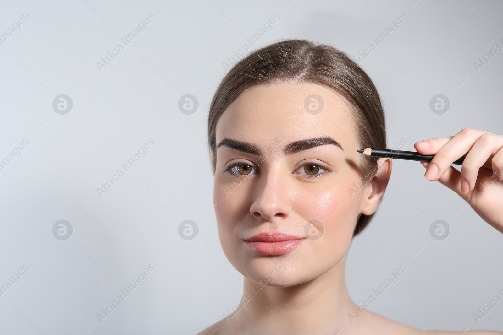 Photo of Young woman correcting shape of eyebrow with pencil on light background