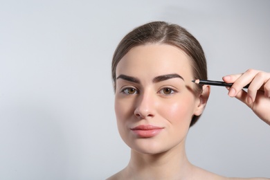 Young woman correcting shape of eyebrow with pencil on light background