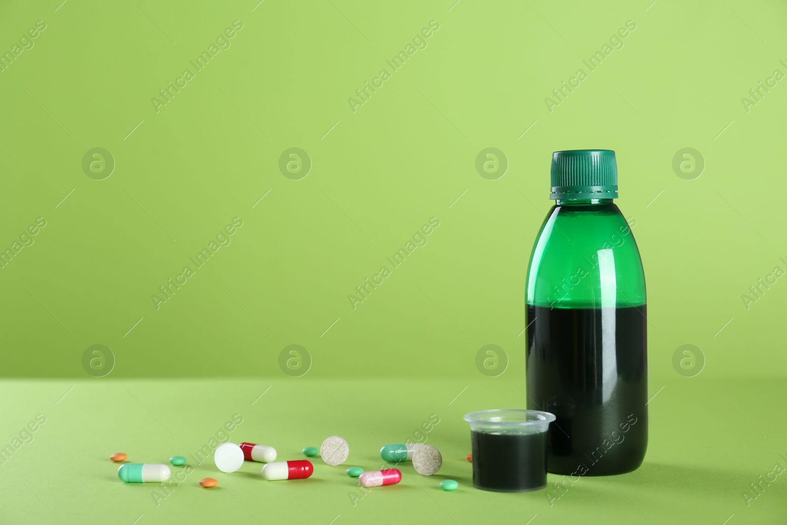 Photo of Bottle of cough syrup, measuring cup and pills on light green background. Space for text